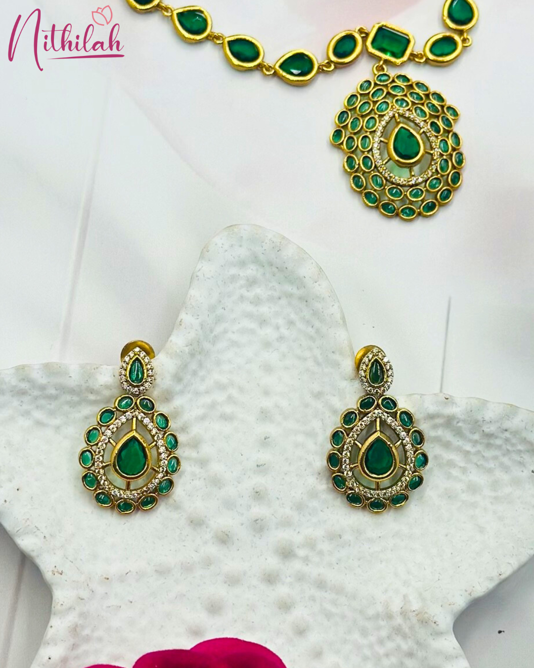 Close-up of gold-finish earrings with emerald-green teardrop centres, oval green stone borders and clear stone accents
