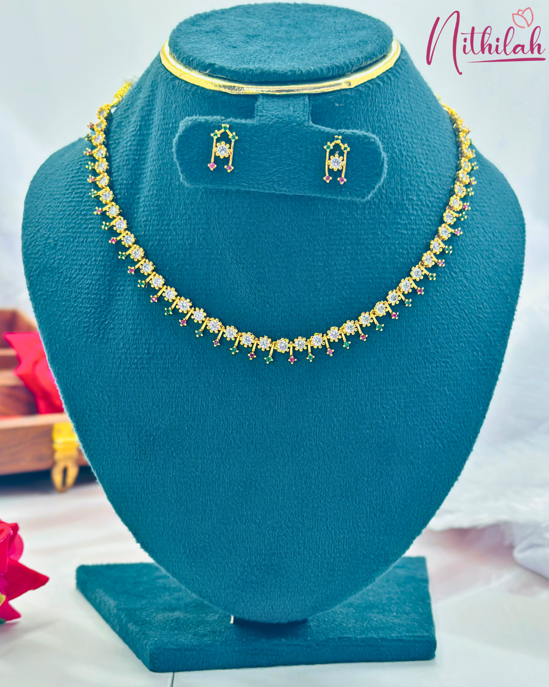 Delicate gold-finish AD necklace and matching earrings with clear stones and pink and green accents on a blue stand