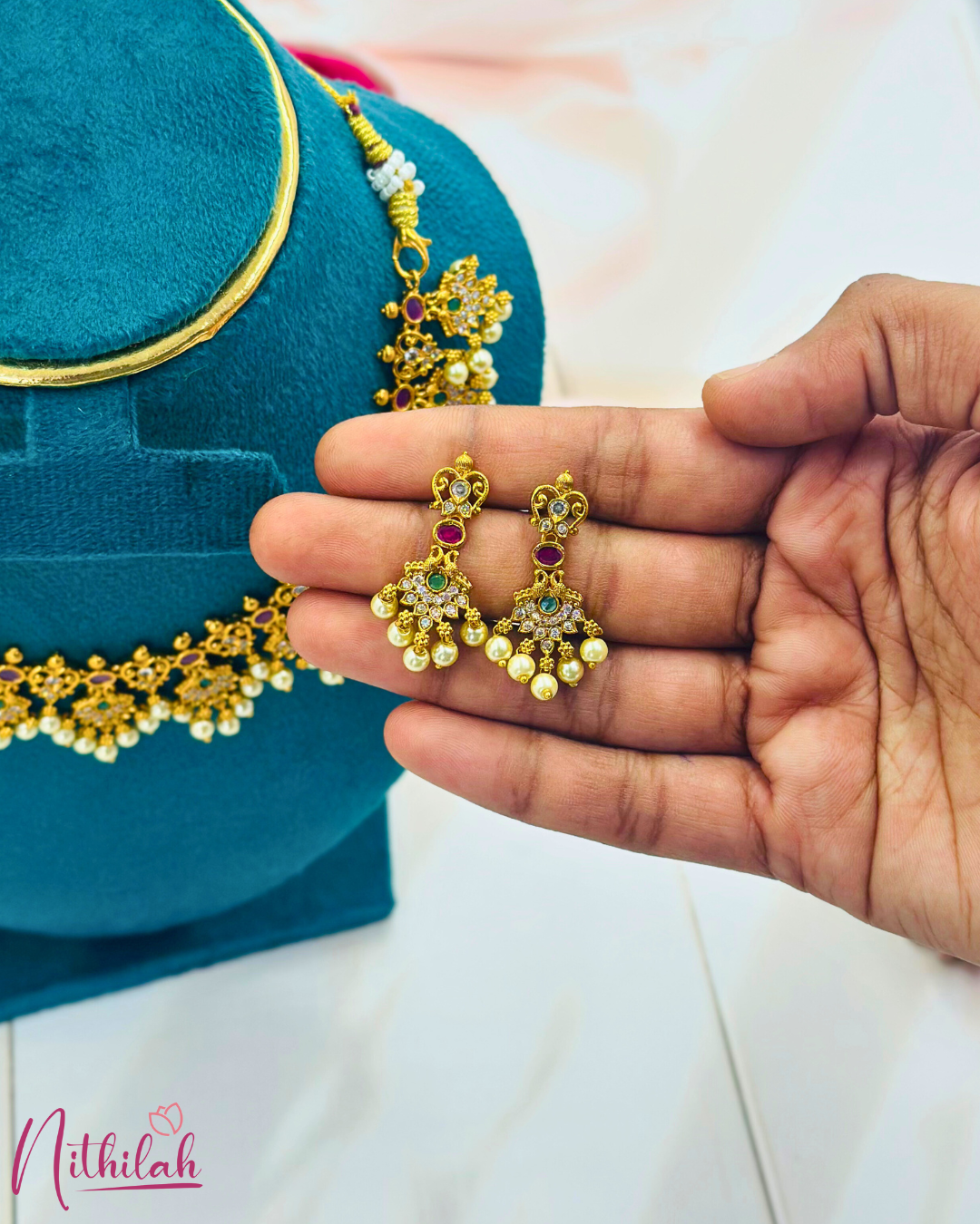 Pair of gold-tone floral drop earrings with purple and green stones and pearl drops, held in a hand