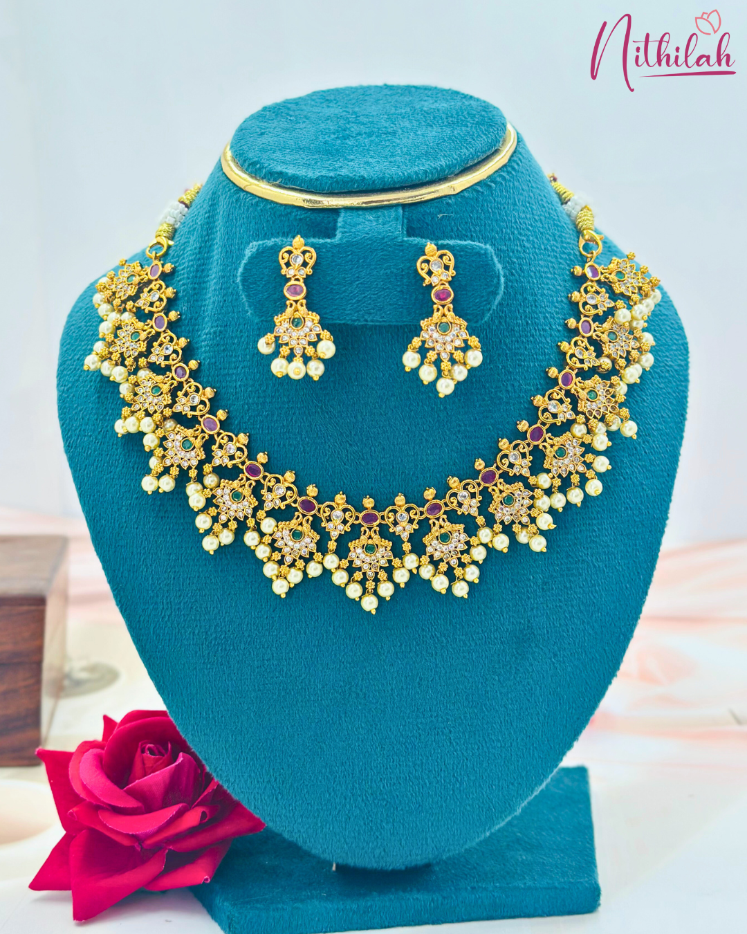 Front view of a gold-tone floral necklace and earring set with purple and green stones and pearl drops on a teal display