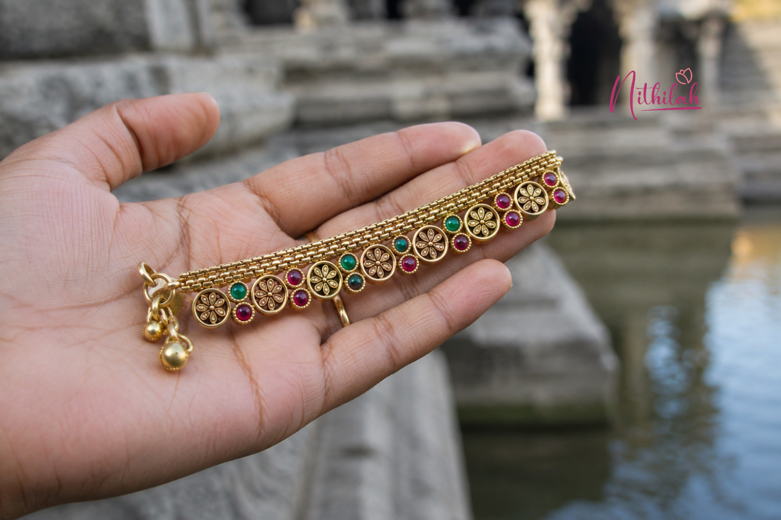 Close-up of an Antique Gold Finish Floral Stone Anklet held in hand near a temple, featuring intricate floral motifs and red-green stone detailing.