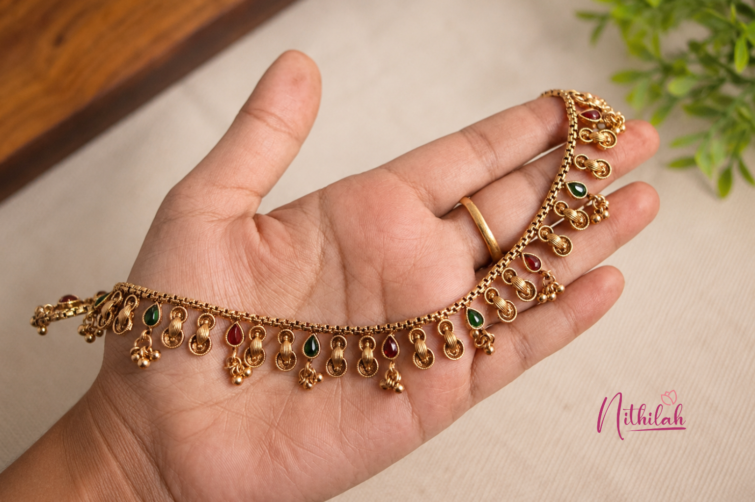 Close-up of an Antique Gold Finish Traditional Anklet with red and green teardrop stones held in hand under natural lighting.