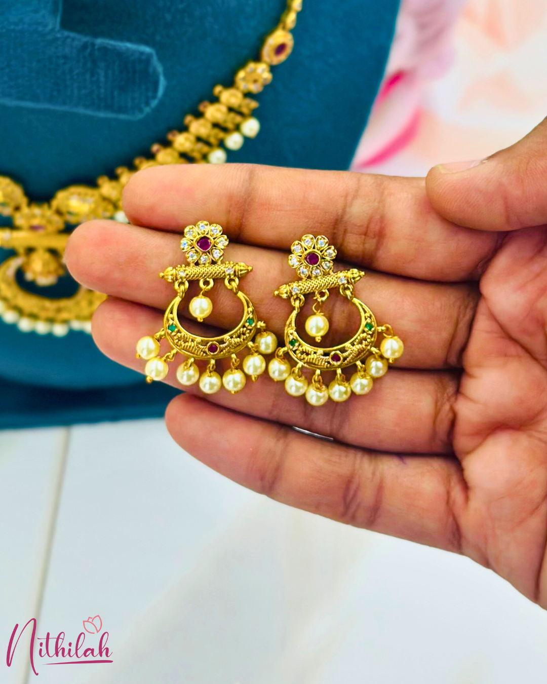 Close-up of matte gold chandbali earrings held in a hand, showing floral studs, coloured stones and pearl drops