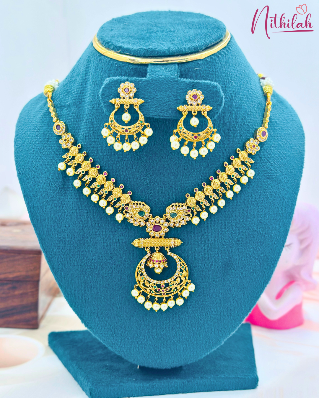 Matte gold peacock necklace and matching pearl-drop earrings displayed on a blue jewellery stand