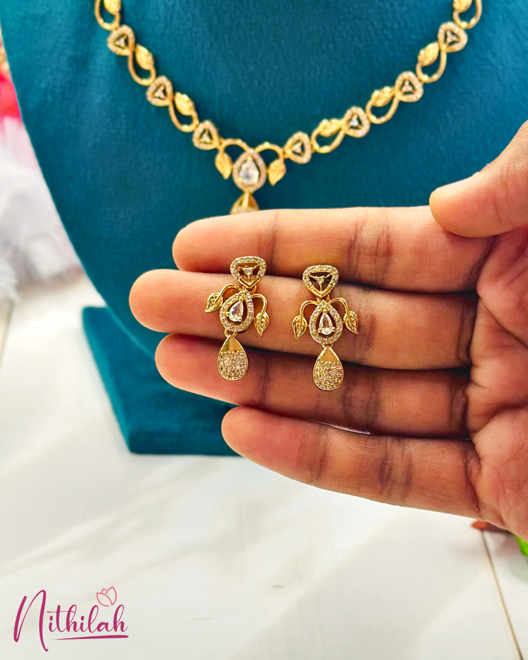 Close-up of gold-plated leaf earrings with clear teardrop stones and CZ-studded drops, held in a hand