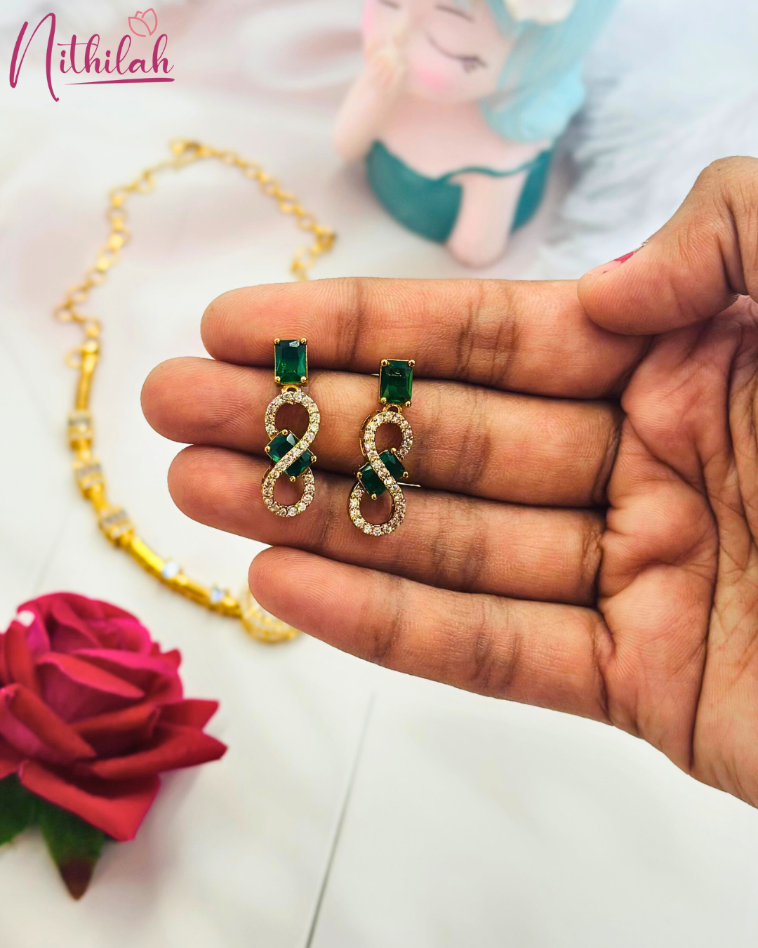 Close-up of gold-plated infinity CZ earrings with rectangular green stones, held in a hand