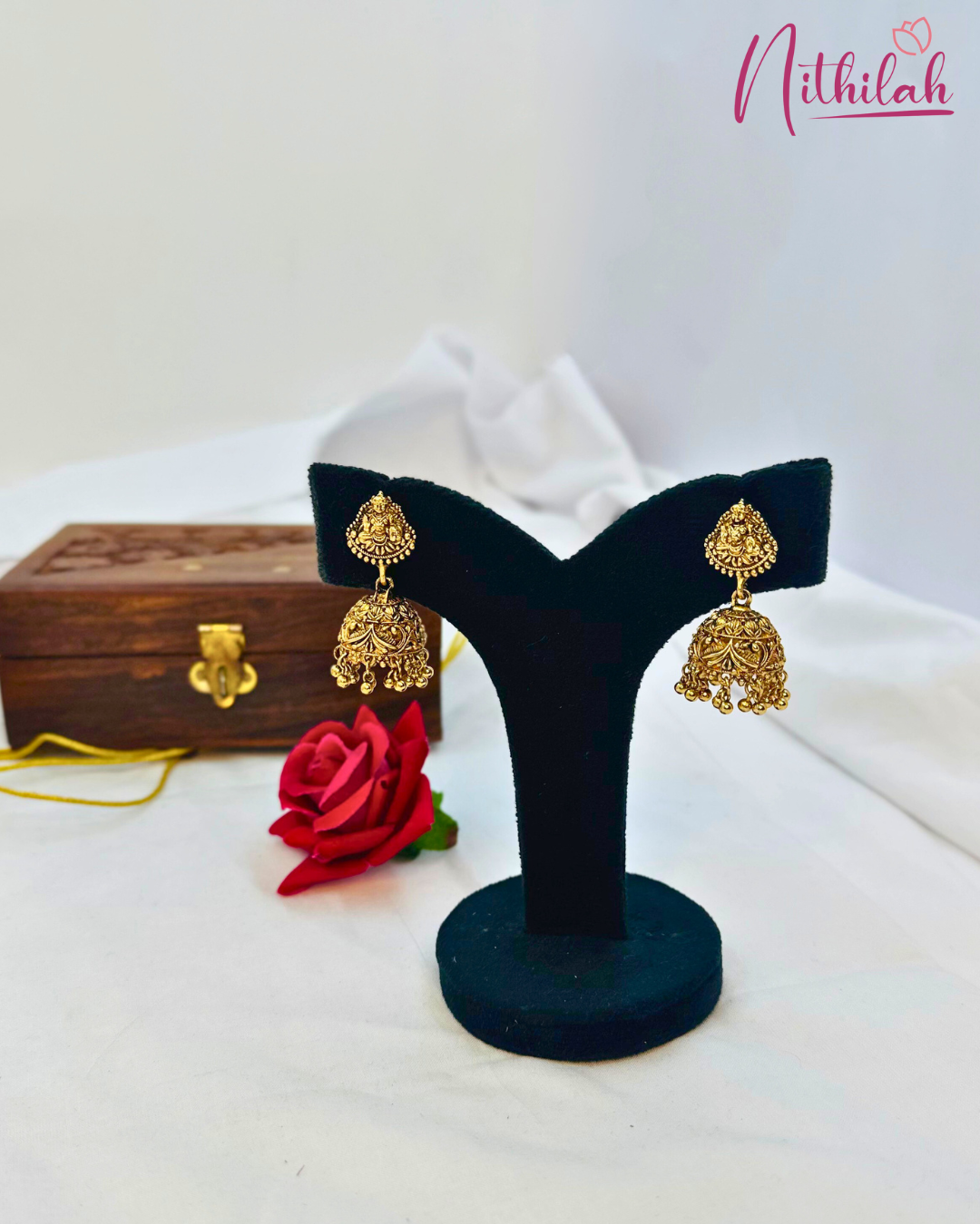 Matte gold-tone jhumka earrings with Lakshmi-motif studs, patterned domes and dangling bead details on a black stand