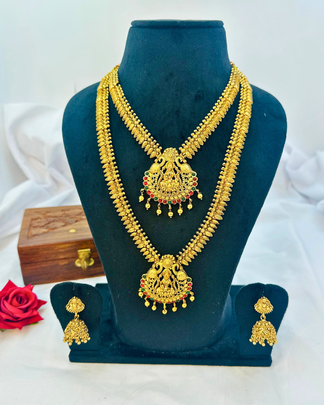 Matte gold-tone Lakshmi haaram and short necklace with red and green pendant accents and matching jhumkas on a display bust