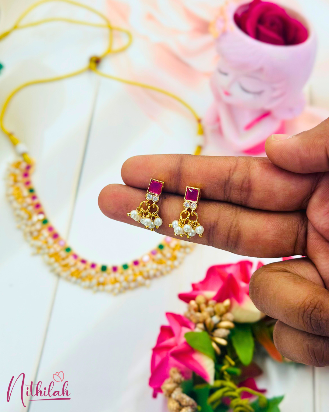Close-up of gold-plated ruby-pink square stone earrings with pearl drops, held in a hand