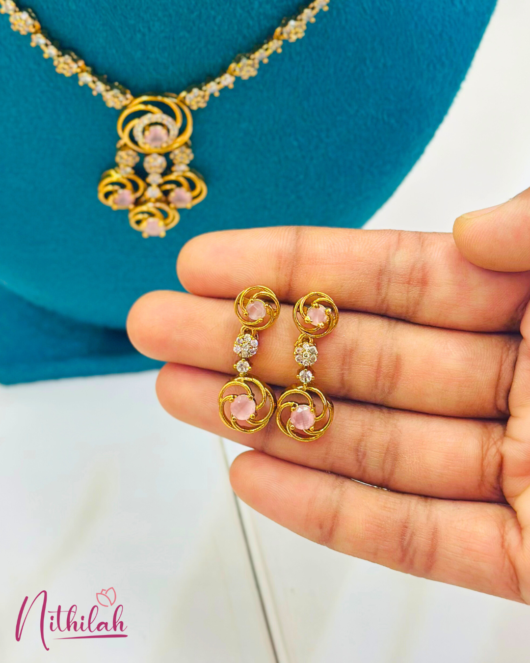 Close-up of pink swirl drop earrings held in a hand, with clear stone links joining gold-finish circular motifs