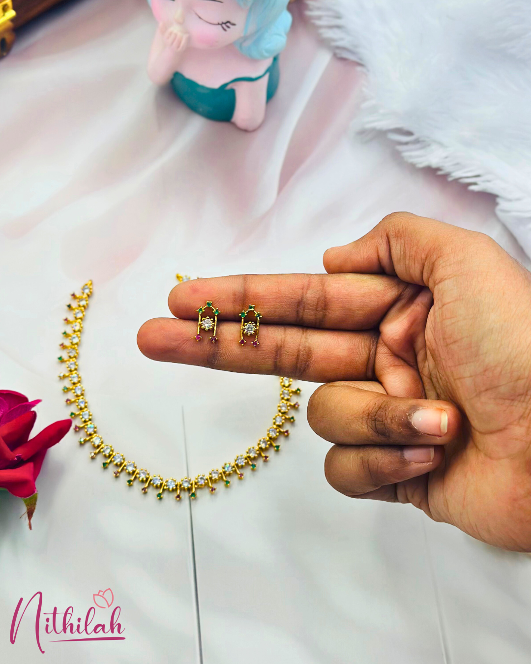 Close-up of small gold-finish earrings held in a hand, showing clear centre stones with pink and green accents