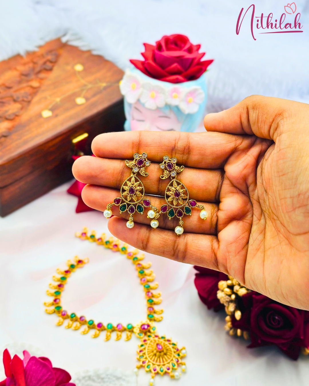 Gold-plated drop earrings held in a hand, showing ruby-pink and green stones, sunburst-style centres and pearl drops