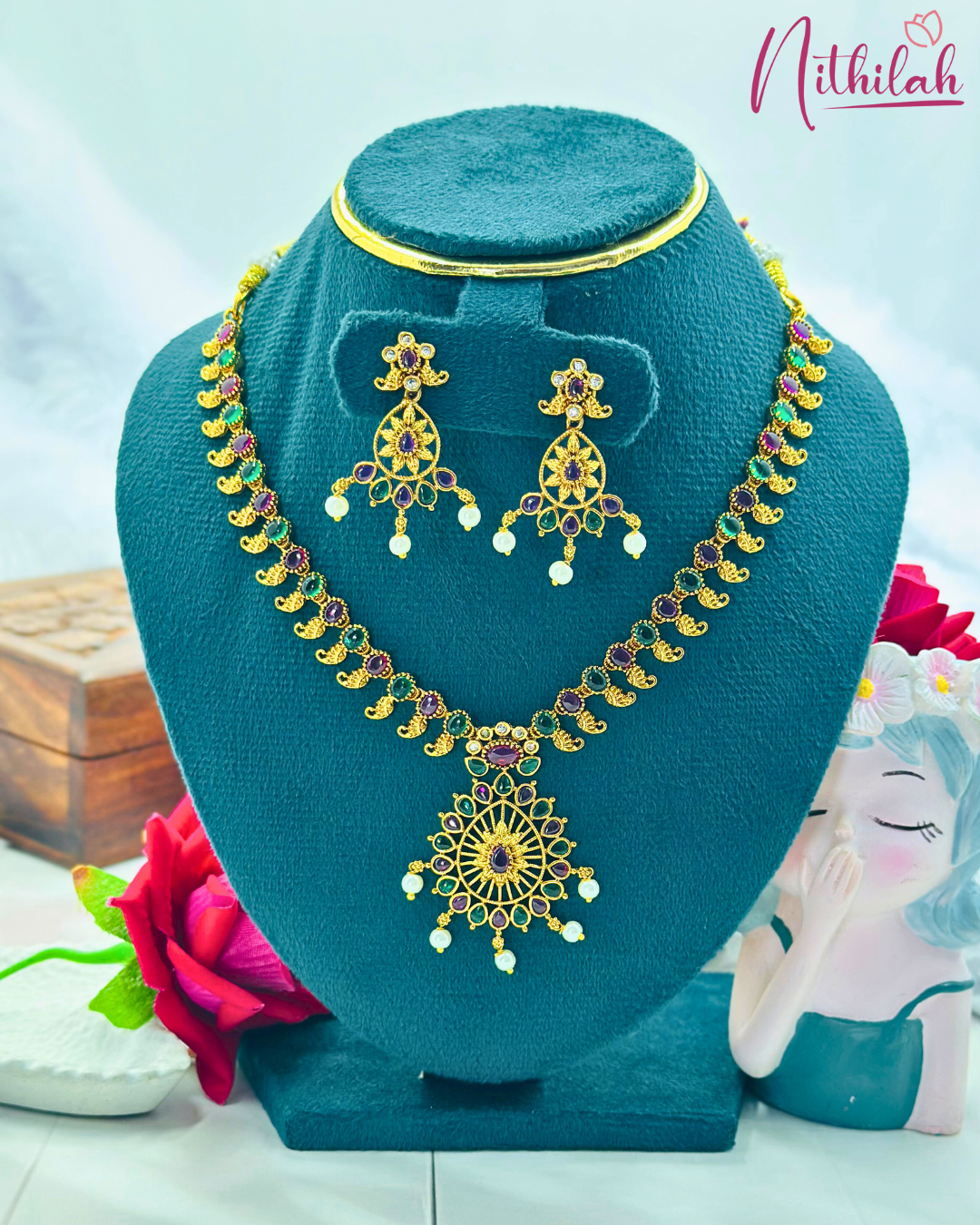 Ruby-pink and green temple-style necklace set with an oval pendant and pearl-drop earrings on a blue jewellery stand