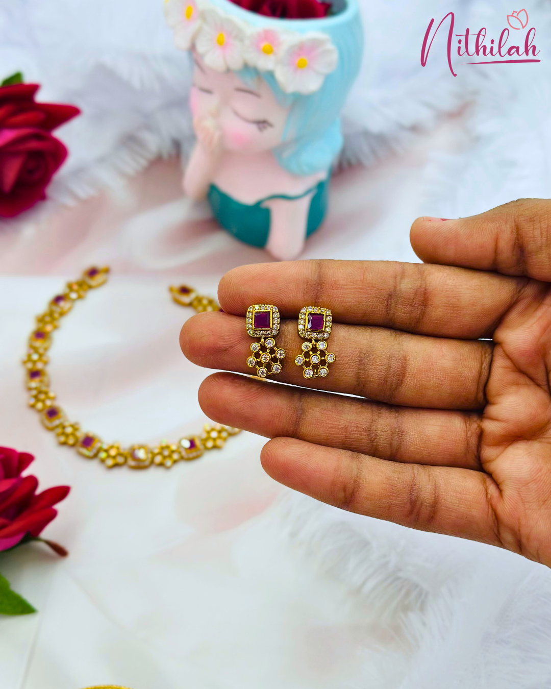 Close-up of gold-finish earrings held in a hand, featuring ruby-pink square stones and clear floral stone drops