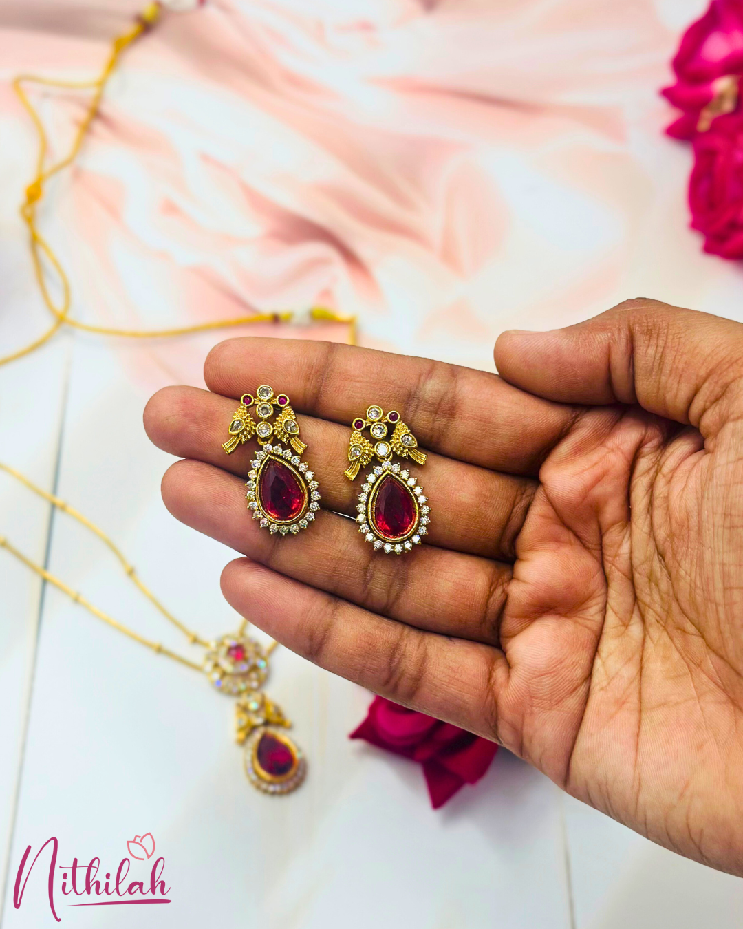 Close-up of matching ruby-pink teardrop earrings held in a hand, with clear stone borders and gold-finish floral detailing