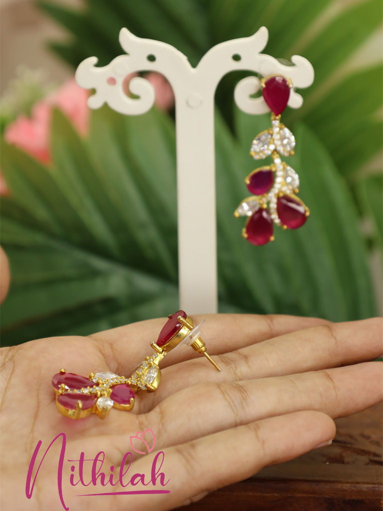 Close-up of ruby red gold-tone drop earrings with clear American Diamond accents, floral leaf detailing, and earring post fastening.